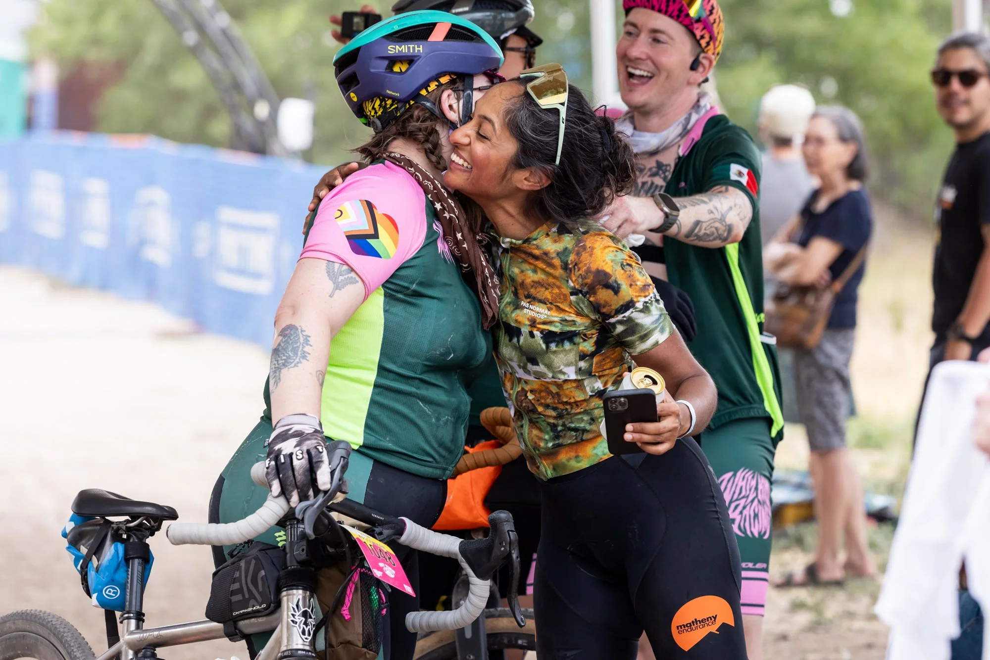 Two women are hugging, one wearing a pink and green cycling shirt with rainbow and heart patches, and the other wearing a colorful camouflage shirt. They are smiling and appear joyful. The woman in camouflage is holding a phone. They are part of a group with others in the background, some wearing helmets and casual clothes, at an outdoor event. The environment is green and appears to be a park or outdoor venue.