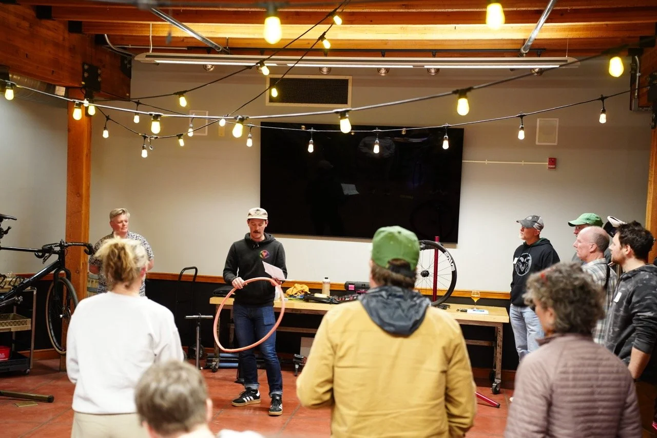 Group of people gathered indoors, listening to a person holding a hula hoop, with bicycles and equipment on tables, in a room with wooden ceiling beams and string lights.