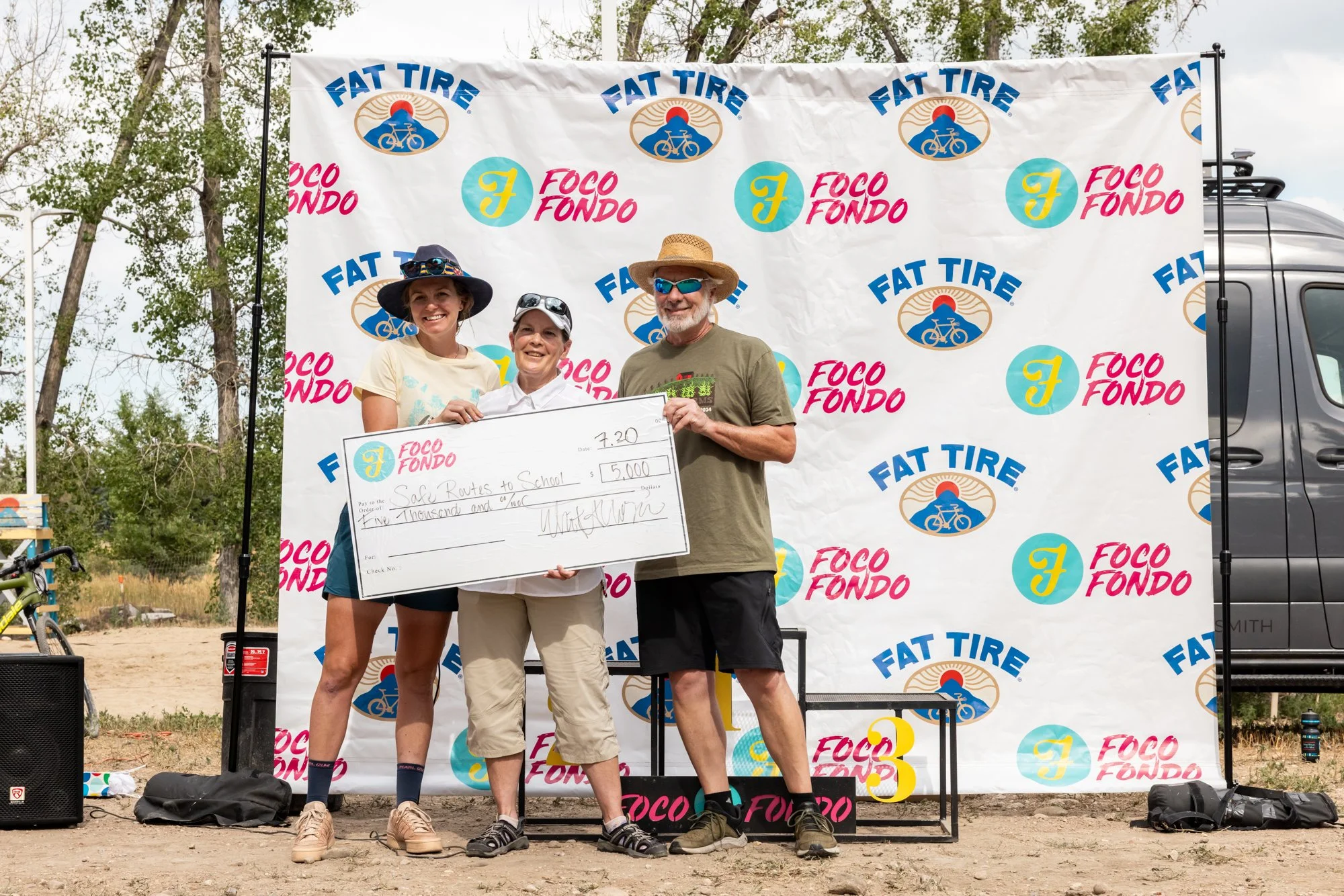 Three people stand on a stage holding a large check at an outdoor event. The backdrop features logos for Fat Tire, Foco Fondo, and other sponsors. Two women and one man are smiling, with the man wearing sunglasses and a straw hat.