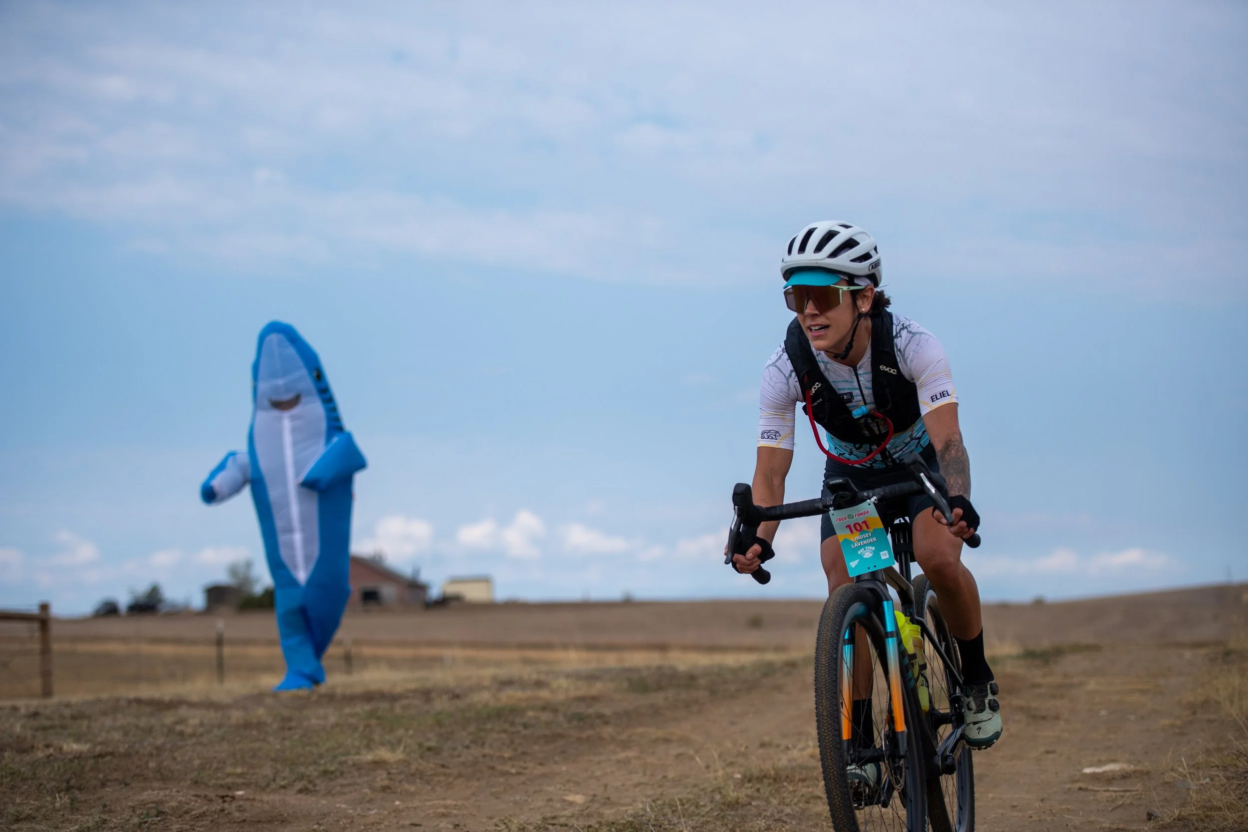 Woman wearing a white cycling helmet and sunglasses riding a mountain bike outdoors on a cloudy day.