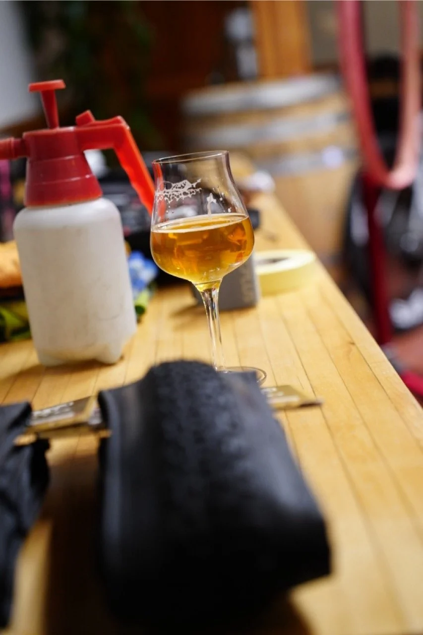 A glass of amber-colored beer on a wooden table with a black leather napkin holder in the foreground.