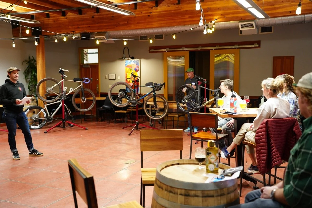 People gathered in a room watching a presentation about bicycles, with bicycle frames mounted on stands and one person speaking.