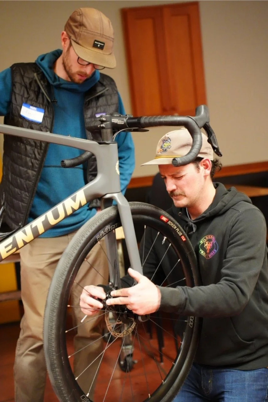 Two men are working on a bicycle indoors. One is standing and looking down at the bike, the other is kneeling and adjusting the bike's rear wheel.