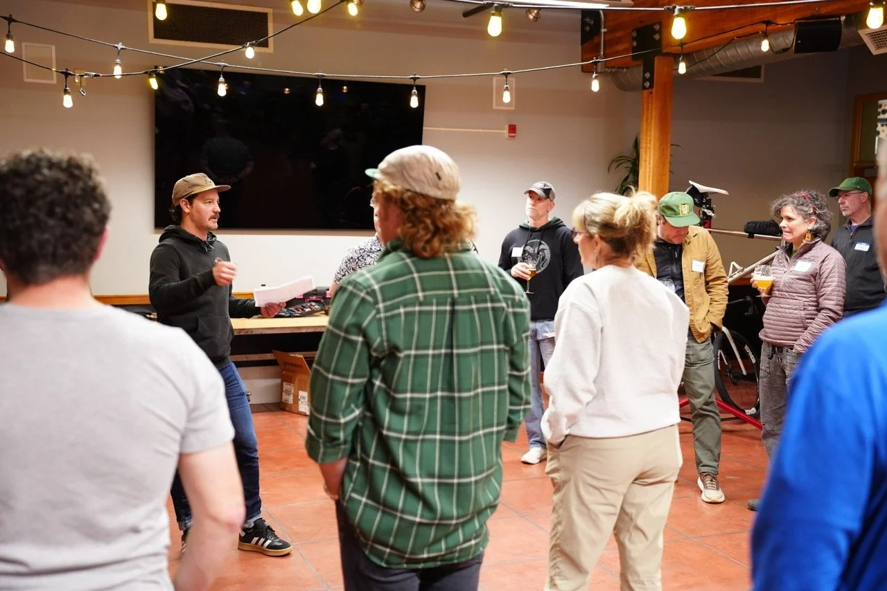 A group of people attending a presentation or workshop in an indoor space, with string lights hanging from the ceiling, and a large black screen or TV on the wall behind the speaker.
