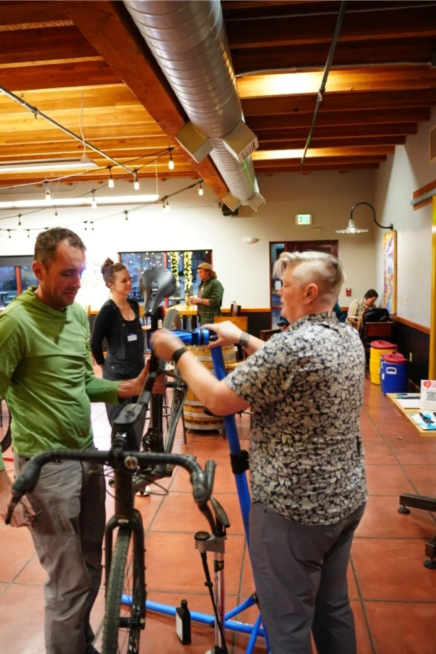 Two men are working together on a bicycle inside a warmly lit indoor space with wooden ceiling beams and string lights. One man is wearing a green shirt, and the other is wearing a patterned shirt.