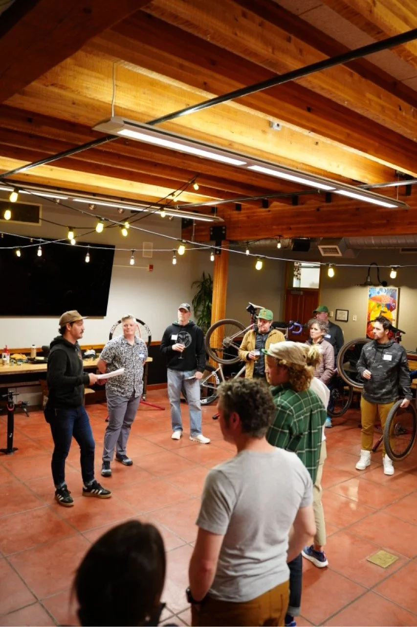 Group of people gathered indoors, some with bicycles, listening to a person reading or speaking, in a room with wooden ceiling beams and string lights.