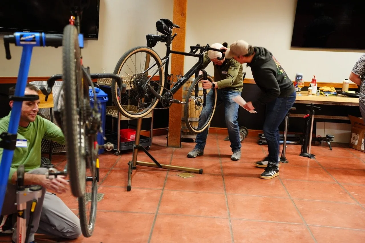 People working on a bicycle mounted on a stand indoors, with one person adjusting the chain and others observing. Various tools and bike parts are visible on the workbench in the background.