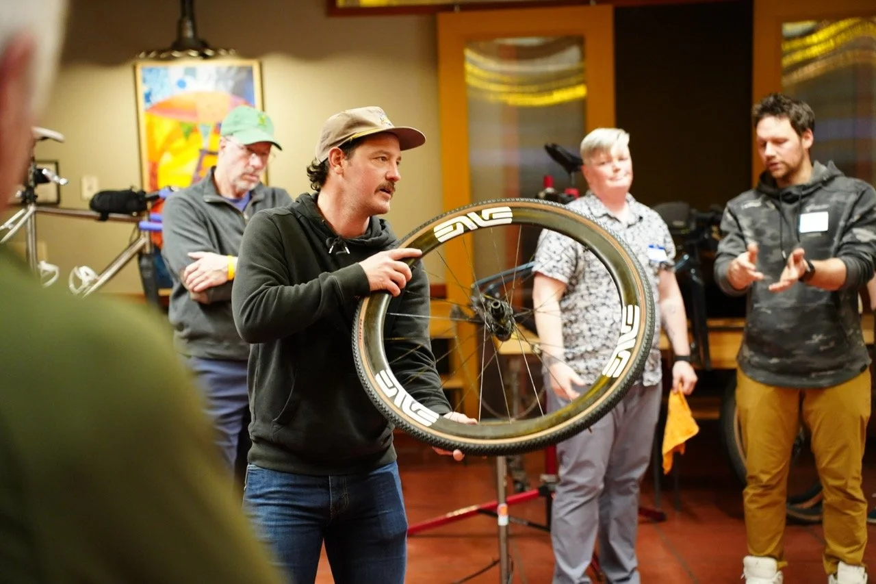 A man demonstrating a bicycle wheel to a group of people indoors, with artwork on the wall behind them.
