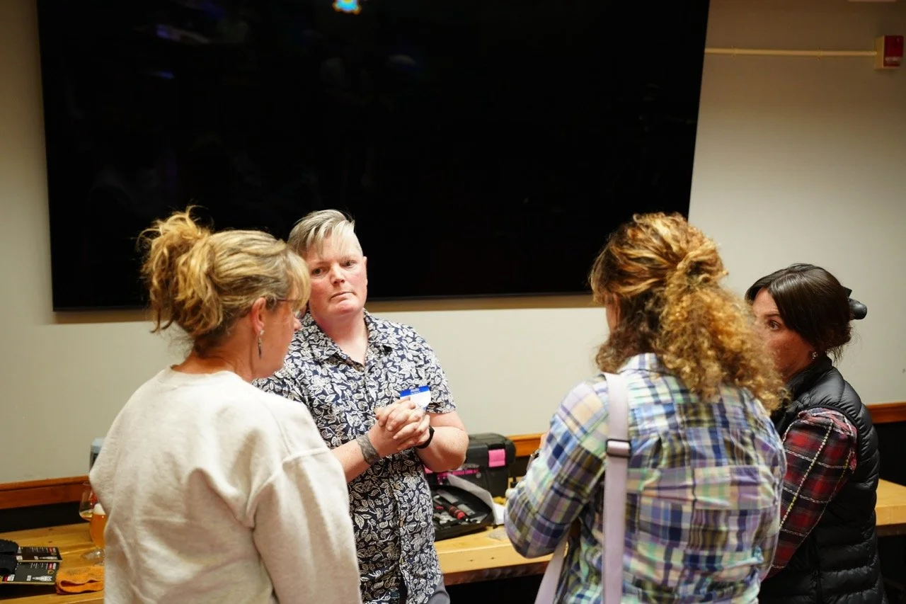 Five women standing and talking in a room with a large black screen on the wall. One woman is holding a small object, and there are various items and tools on the table.