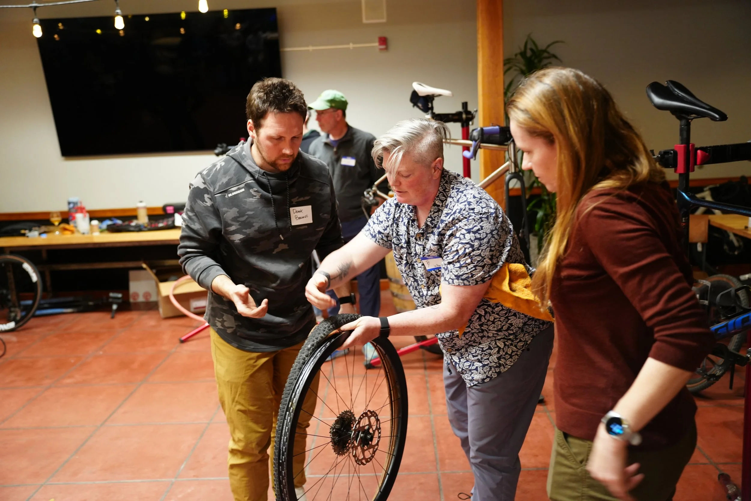 A group of three people in a workshop setting. The person in the middle is holding a bicycle wheel, with a woman on the right looking at it and a man on the left observing. There are tools and a bicycle in the background.