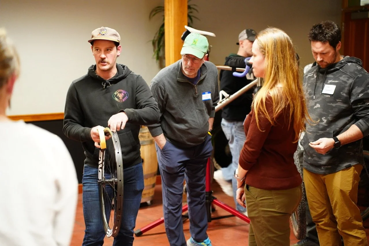Group of people engaged in a discussion indoors, with a man holding a bicycle wheel.