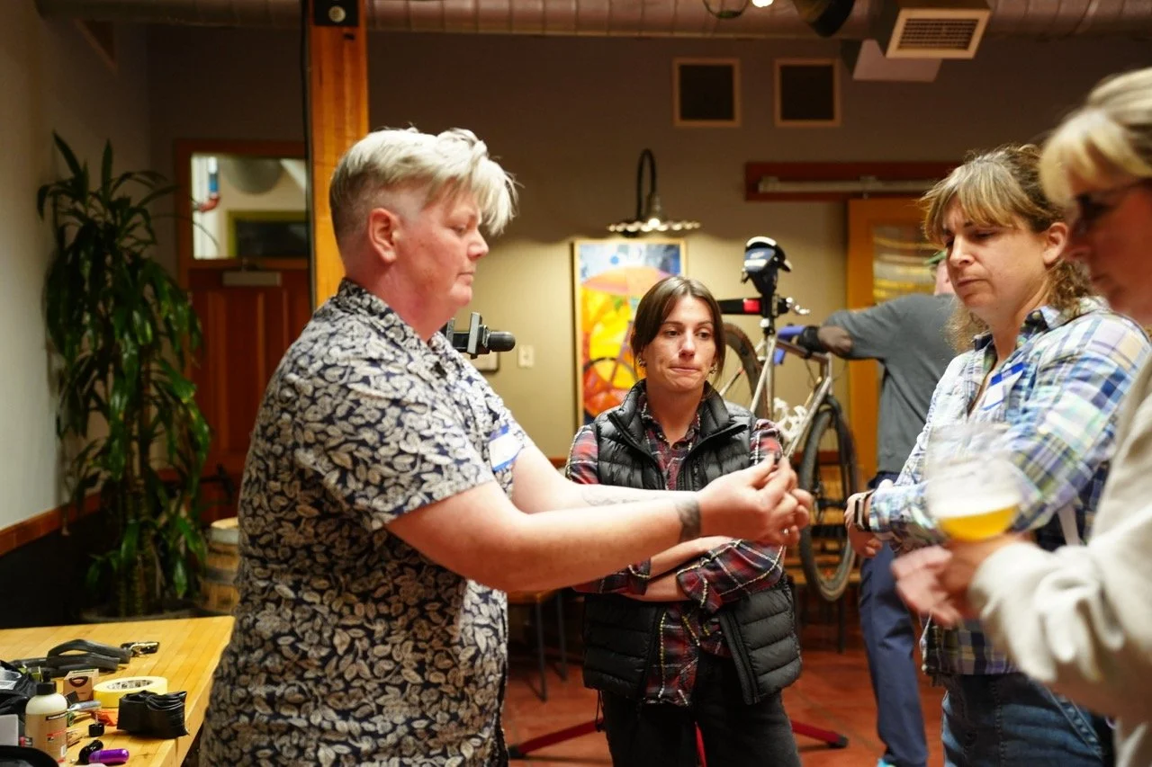 A woman with short blonde hair demonstrating something to a group of women in a workshop or class, with bicycle parts and tools visible on the table, and a bicycle in the background.