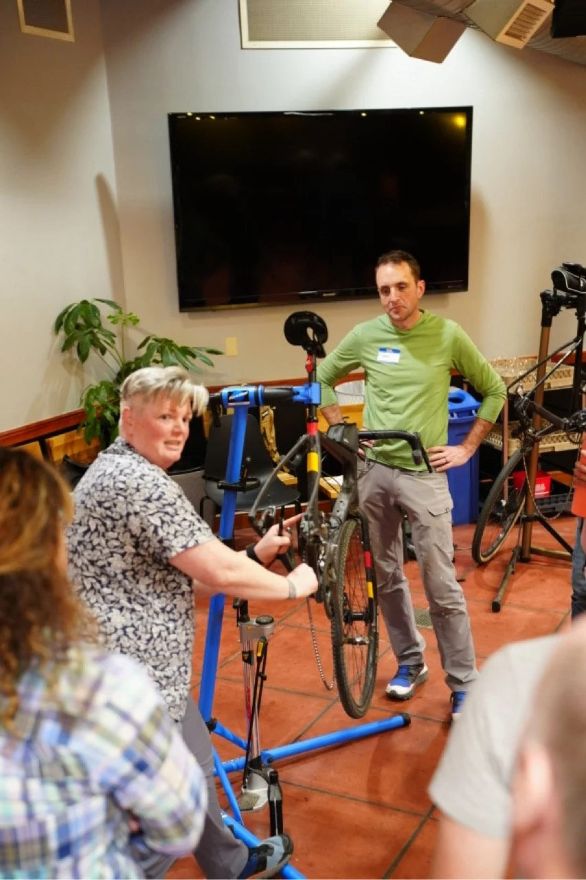 A woman demonstrating bicycle repair or maintenance on a bicycle inside a room. Other people are gathered around watching her. A man in a green shirt looks on, standing with hands on hips. There is a large black television on the wall and some boxes 
