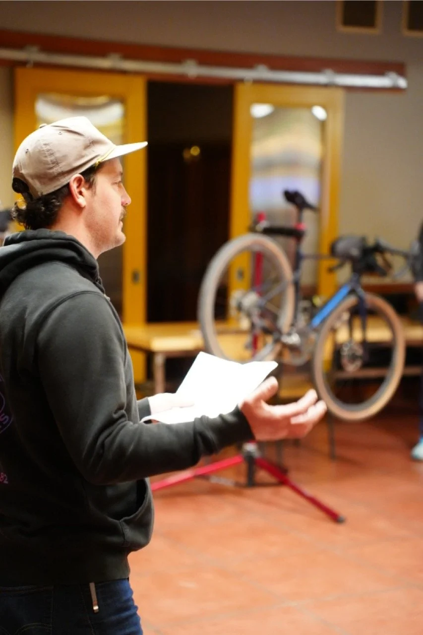 A man wearing a gray hoodie and a beige cap is holding a piece of paper and looking at a bicycle mounted on a stand indoors.