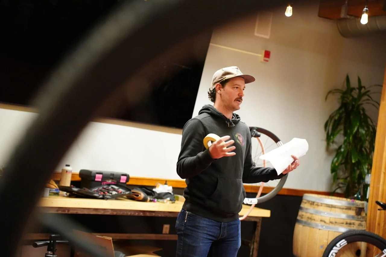 A man wearing a gray cap and a black hoodie stands in a room with wood accents, holding a paper and gesturing as if speaking. Behind him, there is a large television, a wooden barrel, some plants, and tools or equipment on a table.