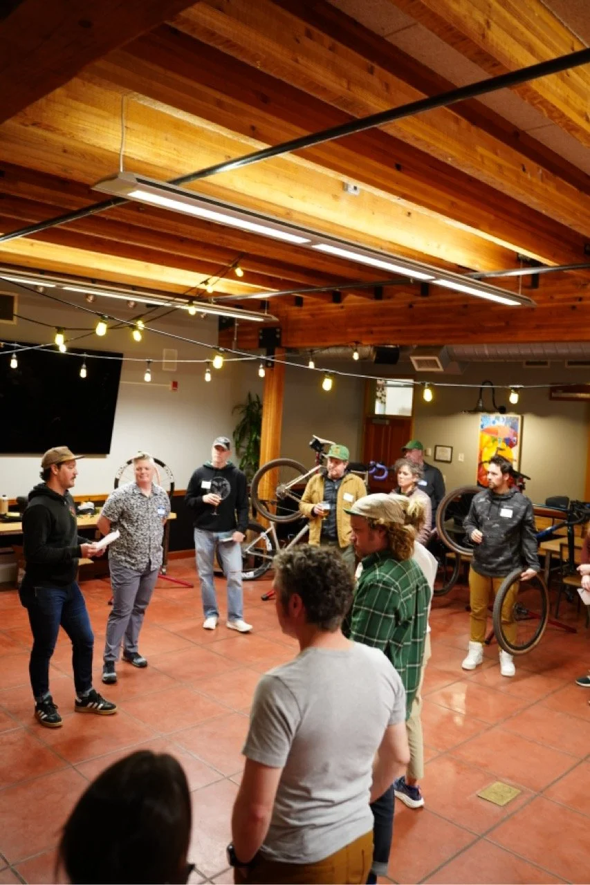 Group of people standing in a circle indoor space with wooden ceiling, string lights, and bicycles in the background, engaging in a conversation or activity.