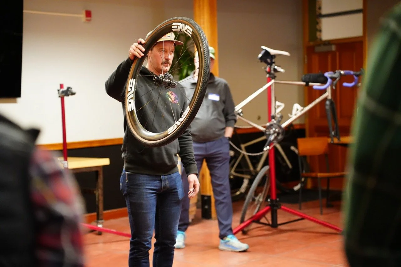 A man holding a bicycle wheel indoors with a bicycle in the background mounted on a stand.