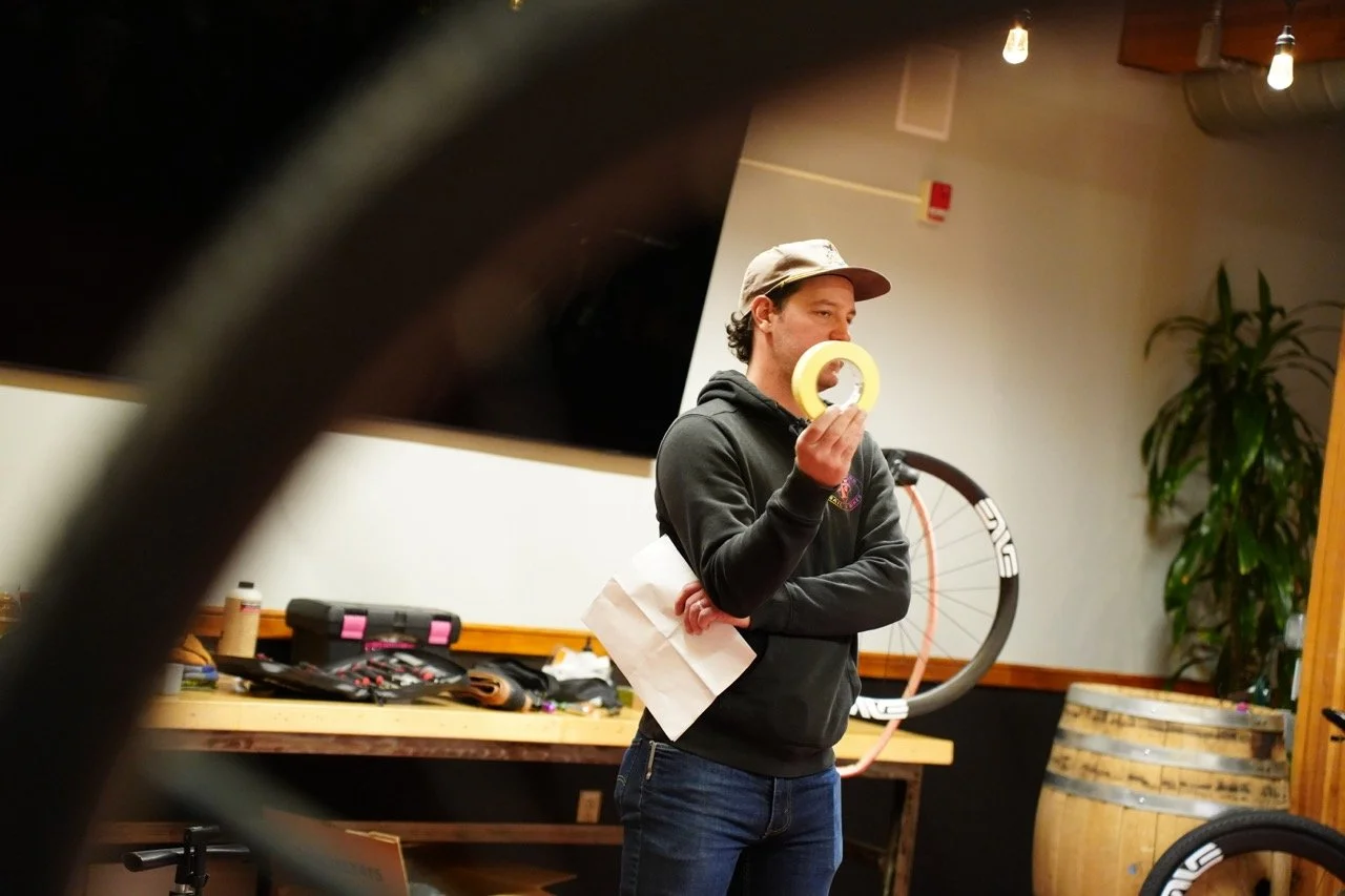 A young man in a black hoodie and gray cap holding a roll of yellow tape in his right hand, standing indoors near a table with tools and papers, with a bicycle wheel behind him and a plant to his right.