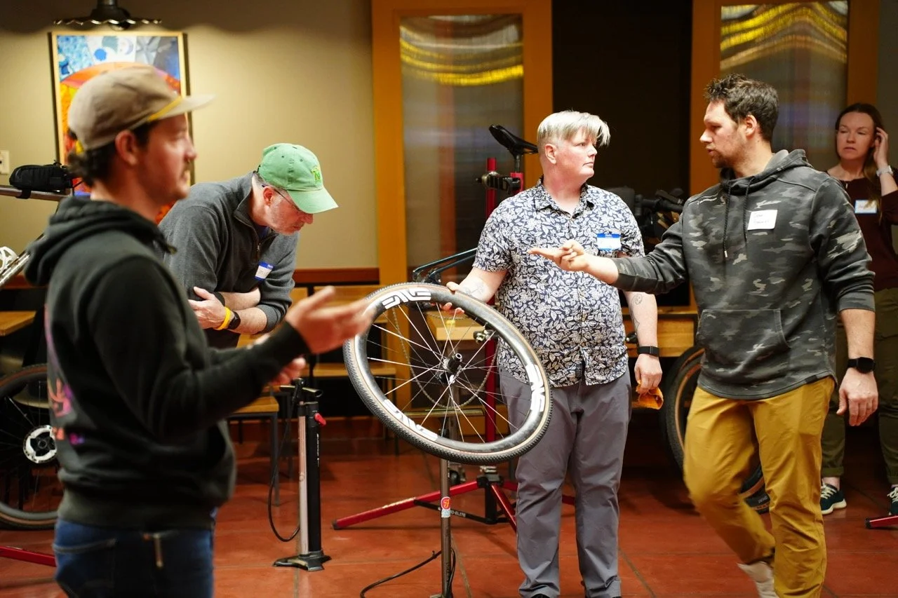 Group of people at a bicycle workshop or class, with one person explaining or demonstrating bike repair or maintenance techniques.