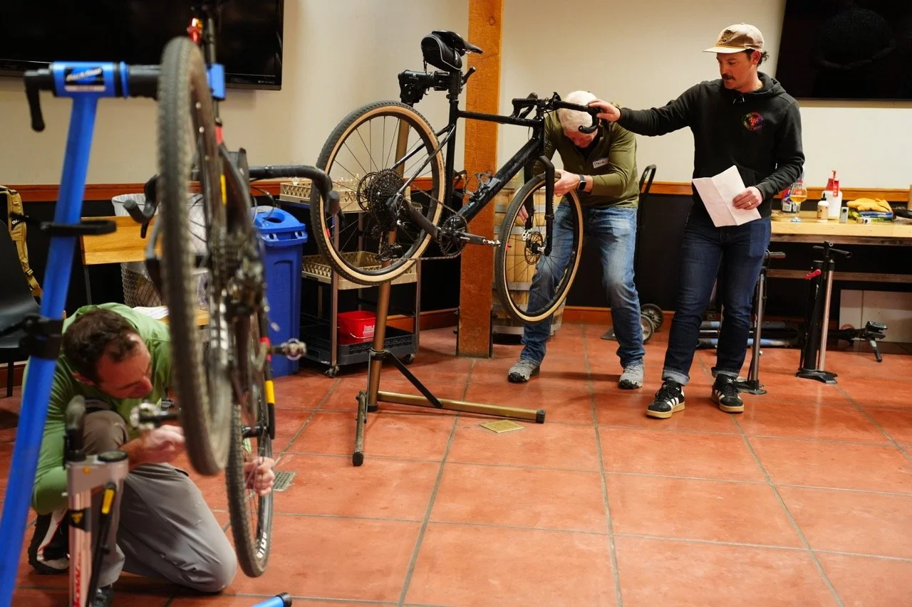 Three men working on a bicycle indoors, with one person holding the bike in the air, another adjusting it, and a third person holding a paper and a pen, on a room with tiled floor and various tools and supplies on a table in the background.