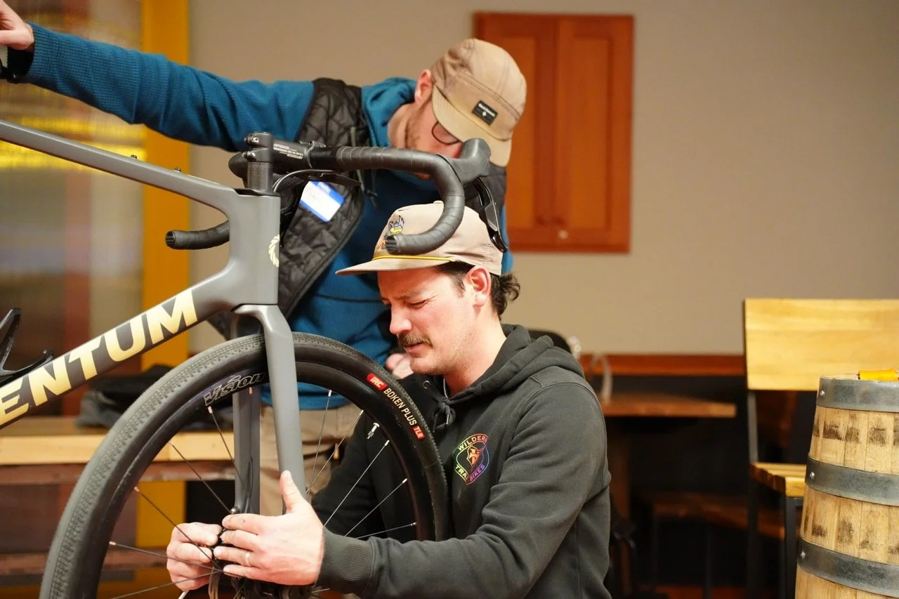 Two men working on a bicycle indoors, one kneeling and the other standing, with a background of wooden furniture and a barrel.