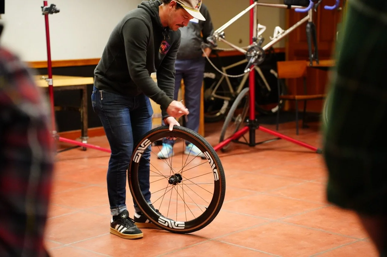 A man in a black hoodie and jeans changing a bicycle wheel indoors with other bicycles and equipment in the background.