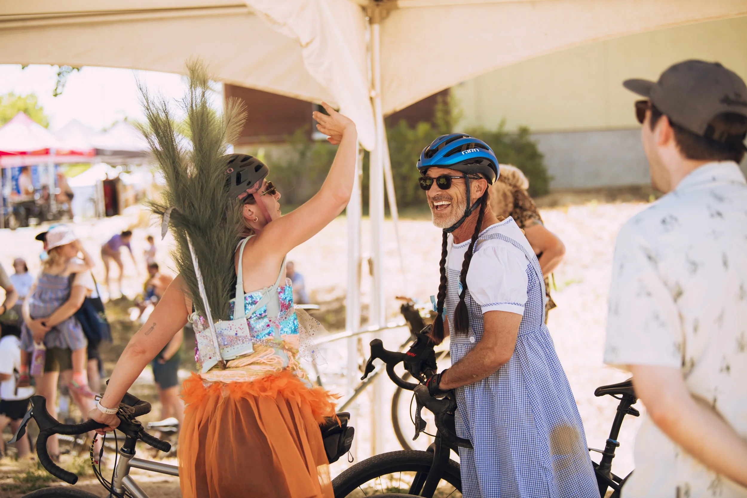 A woman wearing a tutu and a helmet high-fives a smiling man with a beard, wearing sunglasses and a helmet, who is on a bicycle. They are outdoors with other people and tents in the background.