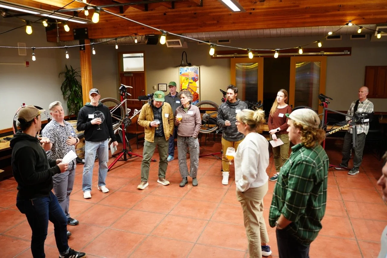 A group of people standing in a semi-circle in a warmly lit indoor space with wooden ceiling, string lights, and bicycles on display in the background. The atmosphere appears casual and conversational, with some holding drinks.