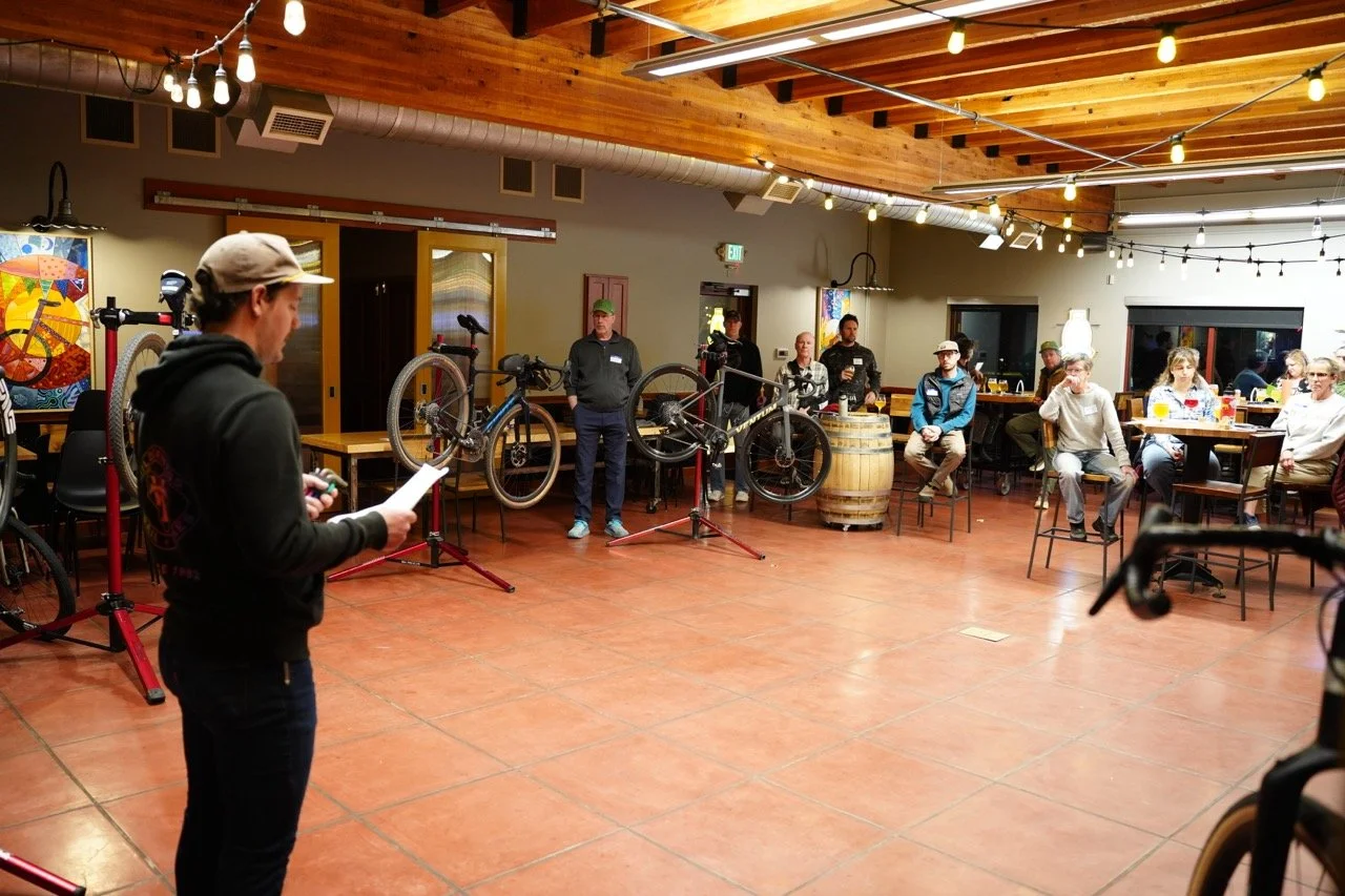 A person is speaking or presenting in front of a group of people at an indoor event, with bicycles and bikes displayed on stands behind them and some people seated at tables in the background.