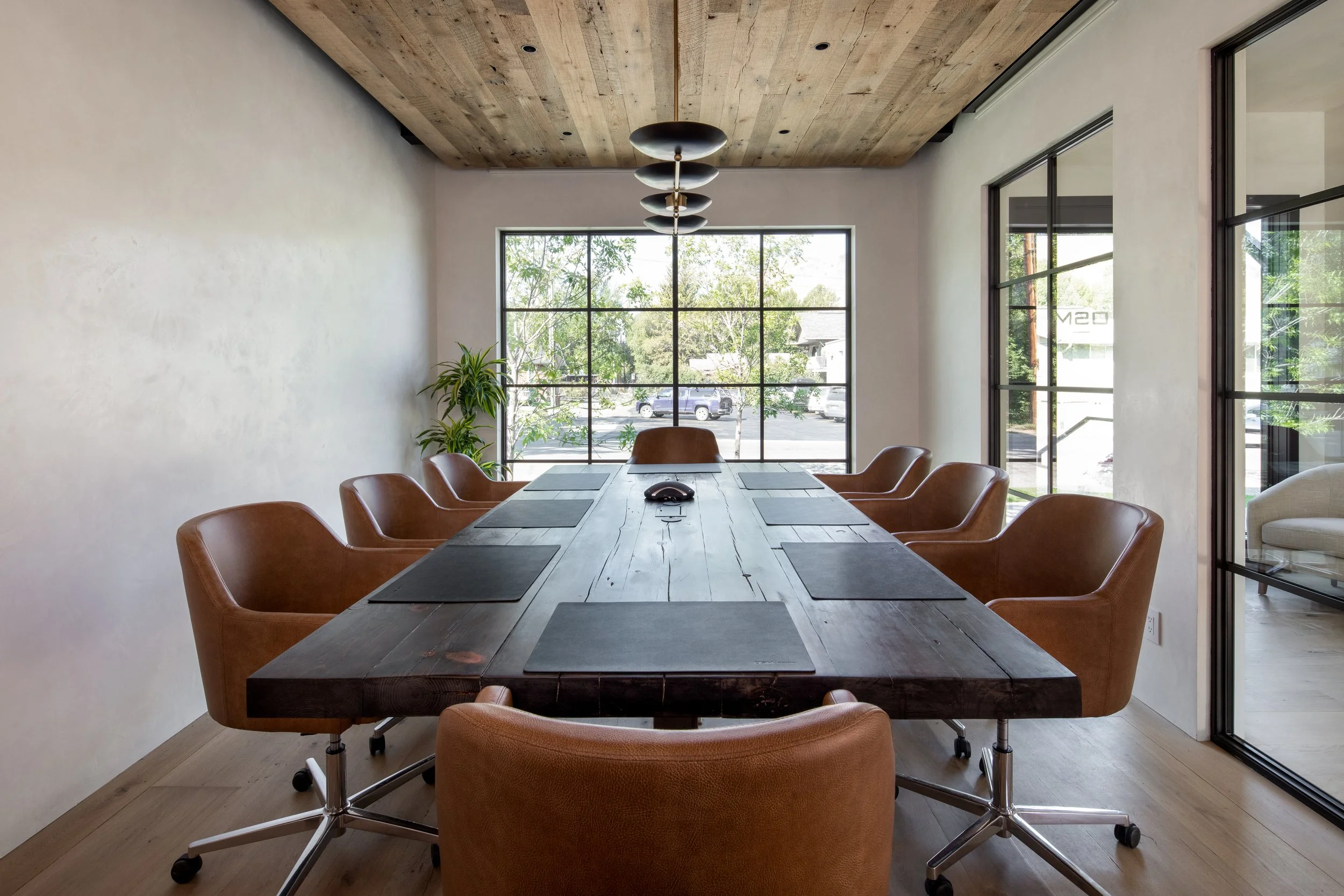 Modern conference room with a long wooden table, black placemats, leather chairs, large windows, and a ceiling light fixture.