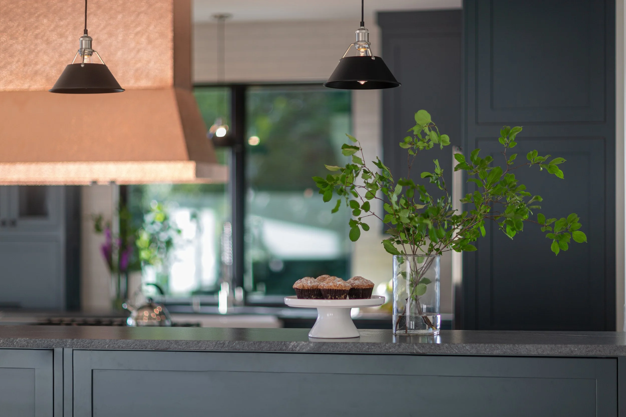 Kitchen counter with a vase of green leafy branches, three muffins on a white stand, and hanging black pendant lights, with a window and cabinets in the background.