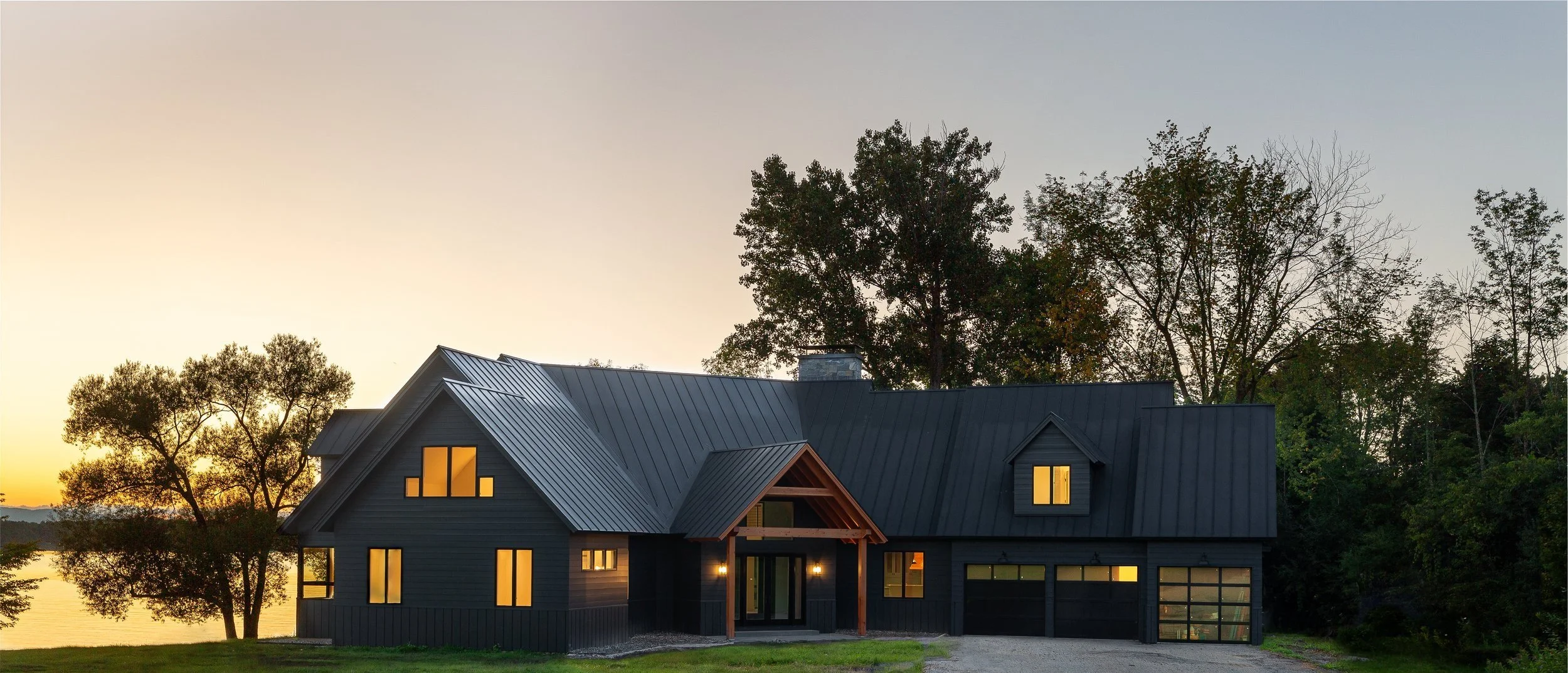 Modern black house with metal roof near water, surrounded by trees, during sunset, with illuminated windows.