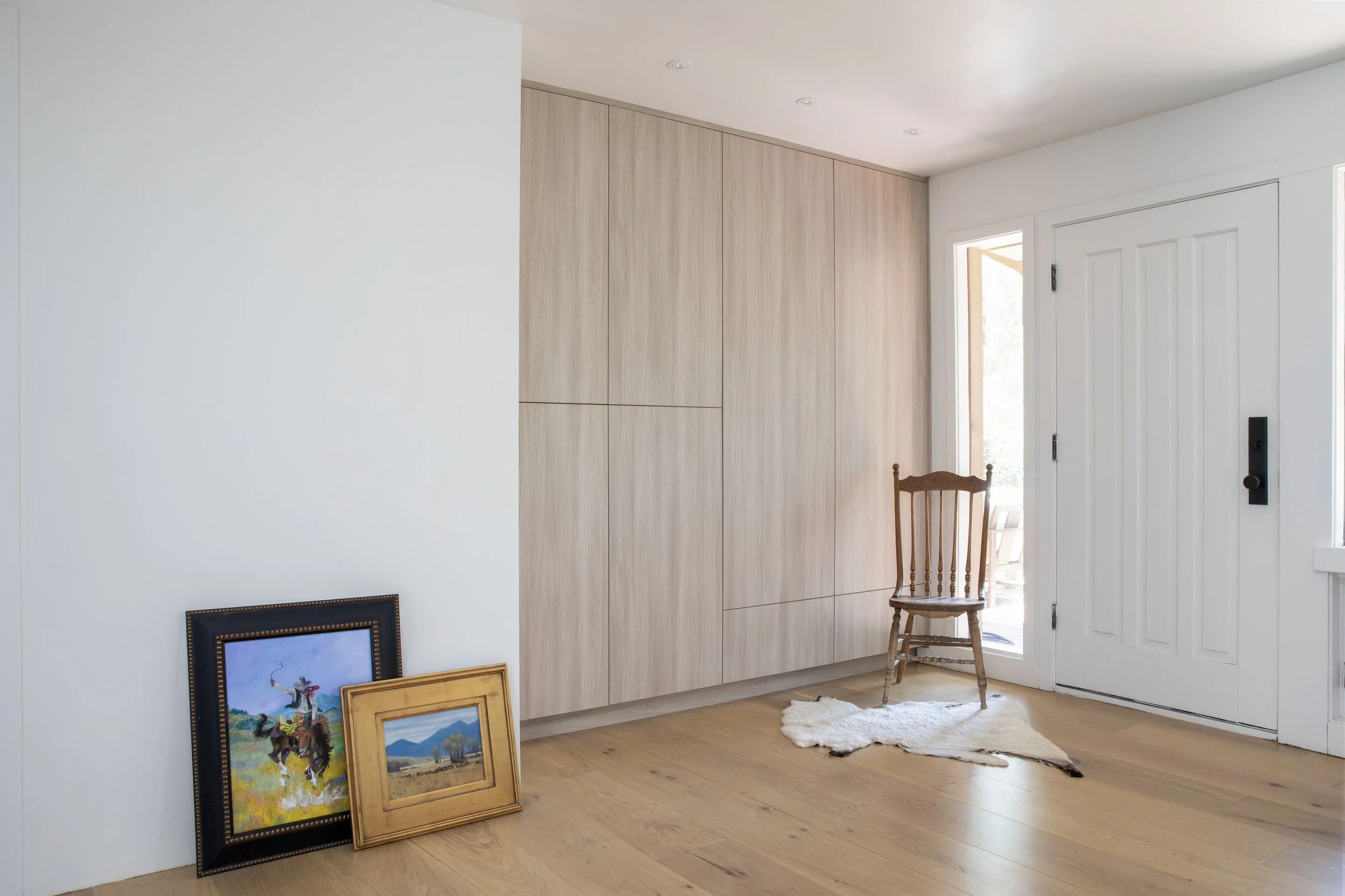 Entryway with a wooden chair on a white sheepskin rug, light wood flooring, paintings leaning against wall near a white door with a black handle, and a built-in light wood closet.