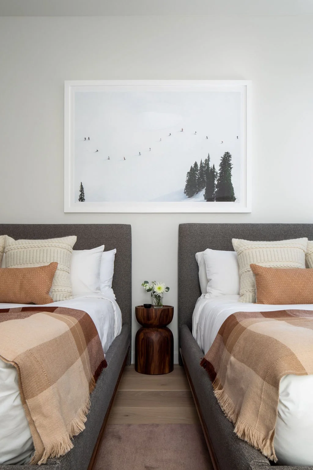 A bedroom with two beds separated by a small wooden nightstand. Each bed has white sheets, multiple beige and cream-colored pillows, and a plaid beige blanket. There is a framed mountain landscape photograph hanging on the wall above the beds, and a small vase with white flowers on the nightstand.