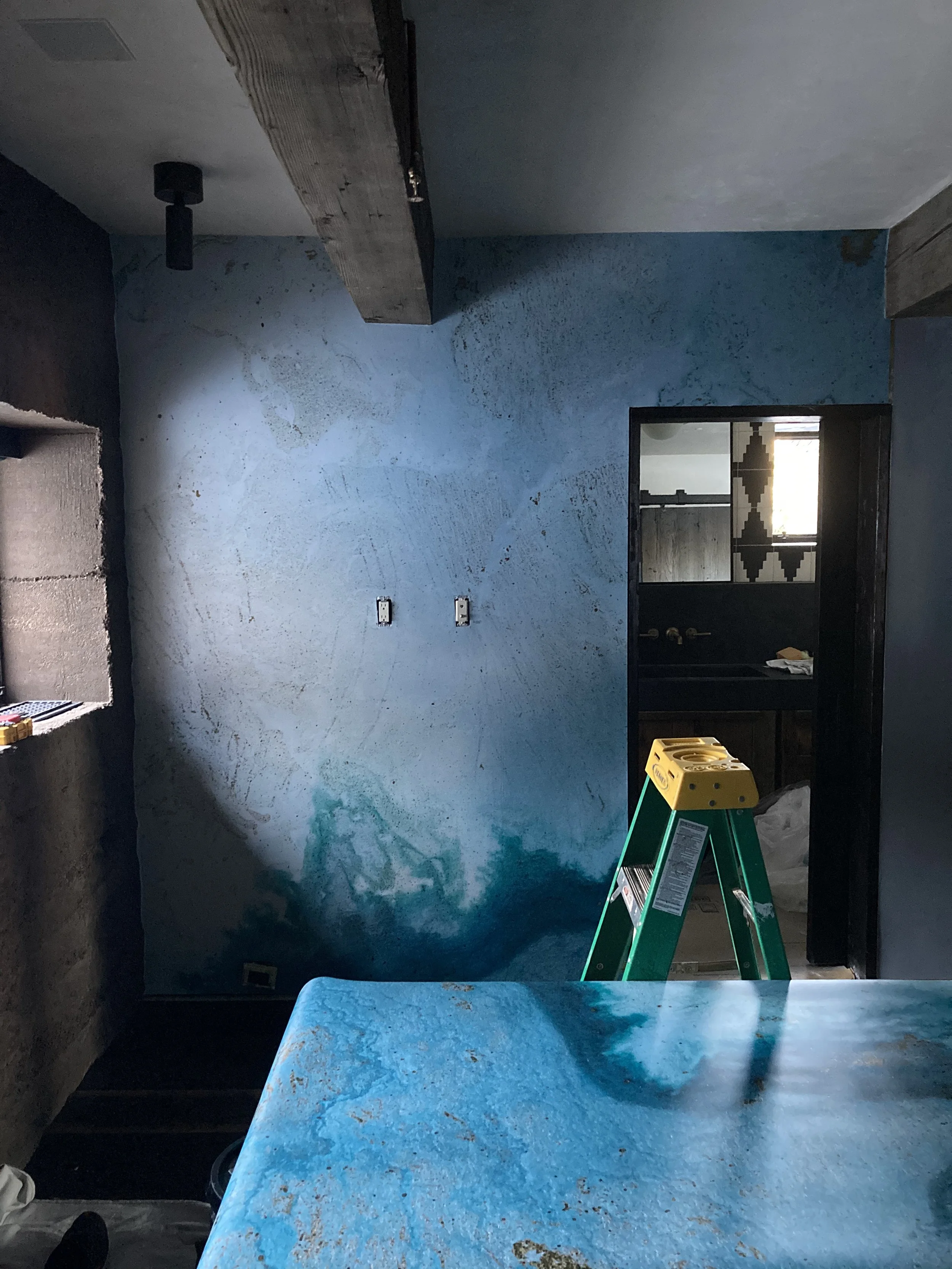 A room under construction or renovation with a blue textured wall, a green ladder, a black doorway, and an unfinished countertop with construction tools and materials.
