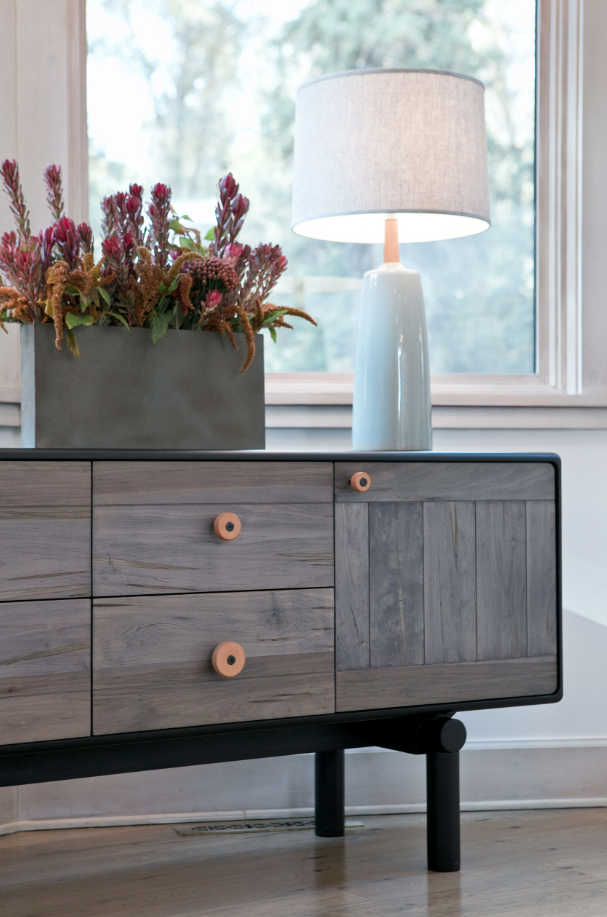 A wooden sideboard with round wooden knobs, topped with a gray rectangular planter filled with pink flowers, and a white ceramic table lamp with a light gray shade, placed in front of a window.