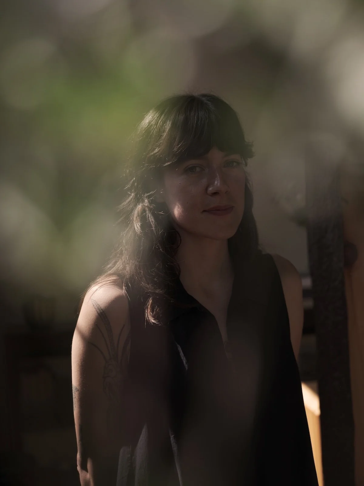 A woman with dark hair and a tattoo on her arm, wearing a sleeveless black top, standing indoors, with soft natural light and blurred surroundings.