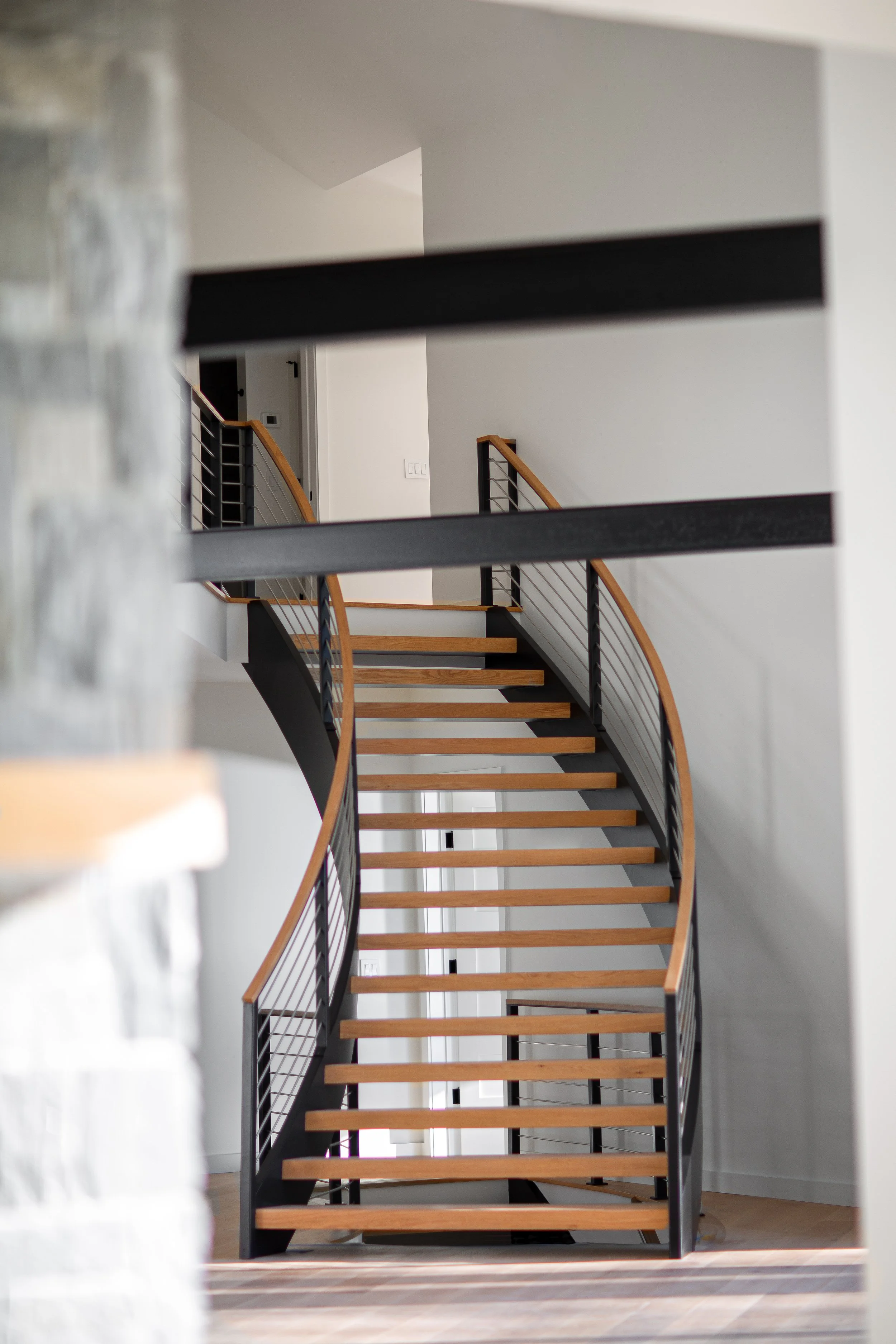 Empty modern staircase with wooden steps and black metal railings, viewed through a partial blurred foreground wall inside a house.