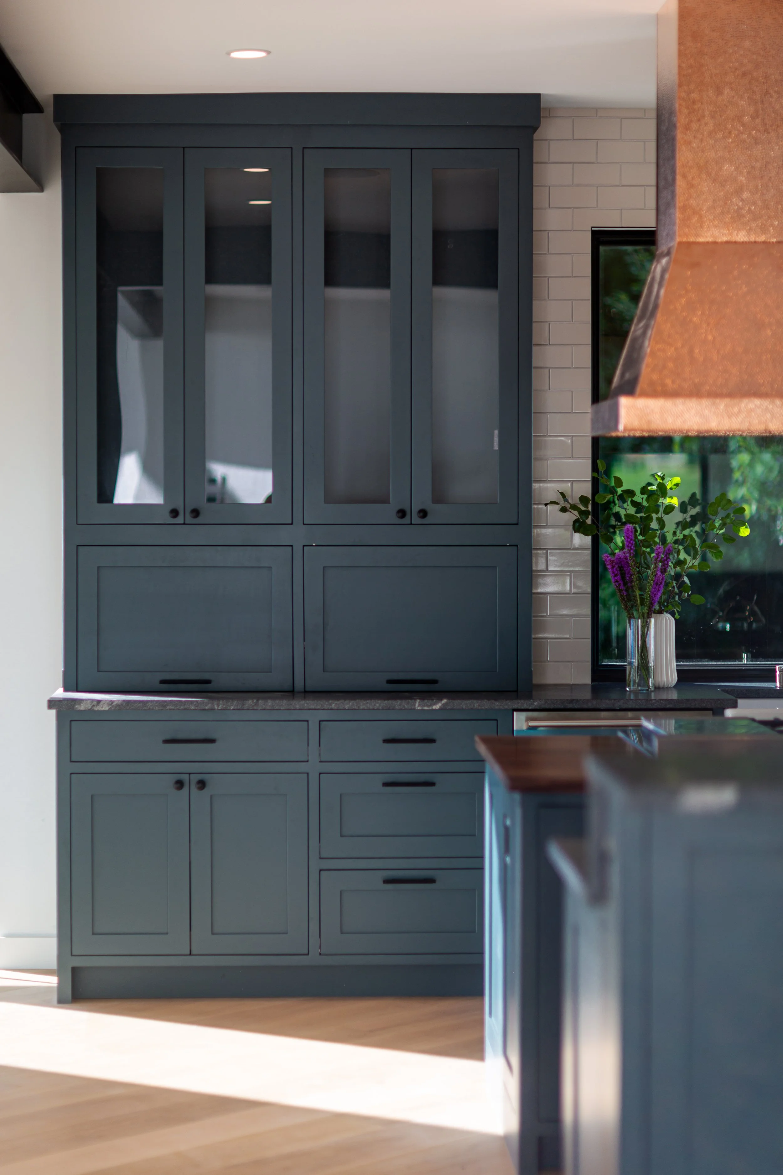 Modern kitchen with gray cabinetry, a dark countertop, a window with green foliage outside, and a vase of purple flowers on the counter.