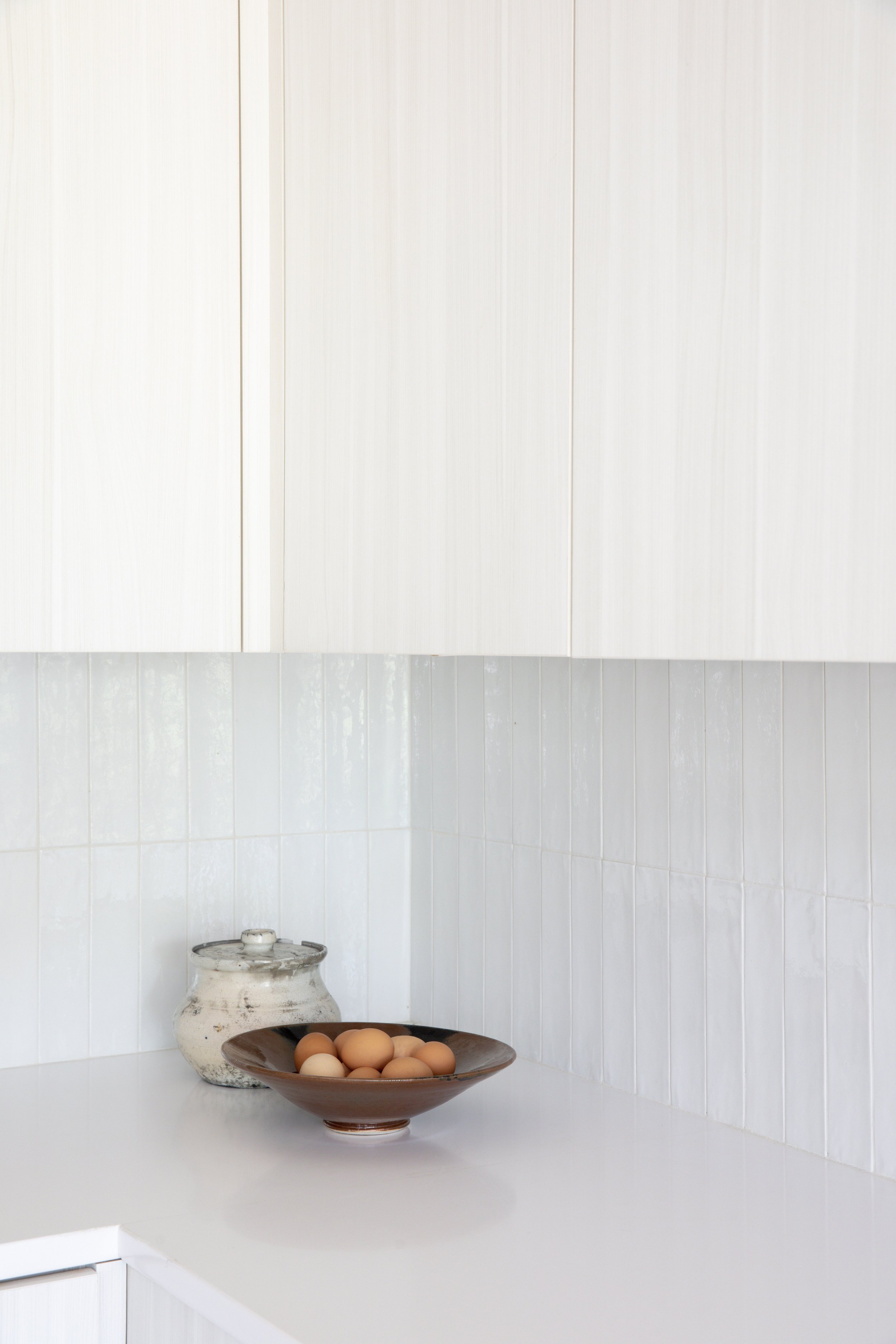 A kitchen countertop with a rustic ceramic jar and a brown bowl filled with eggs, with white cabinets and tiled walls in the background.