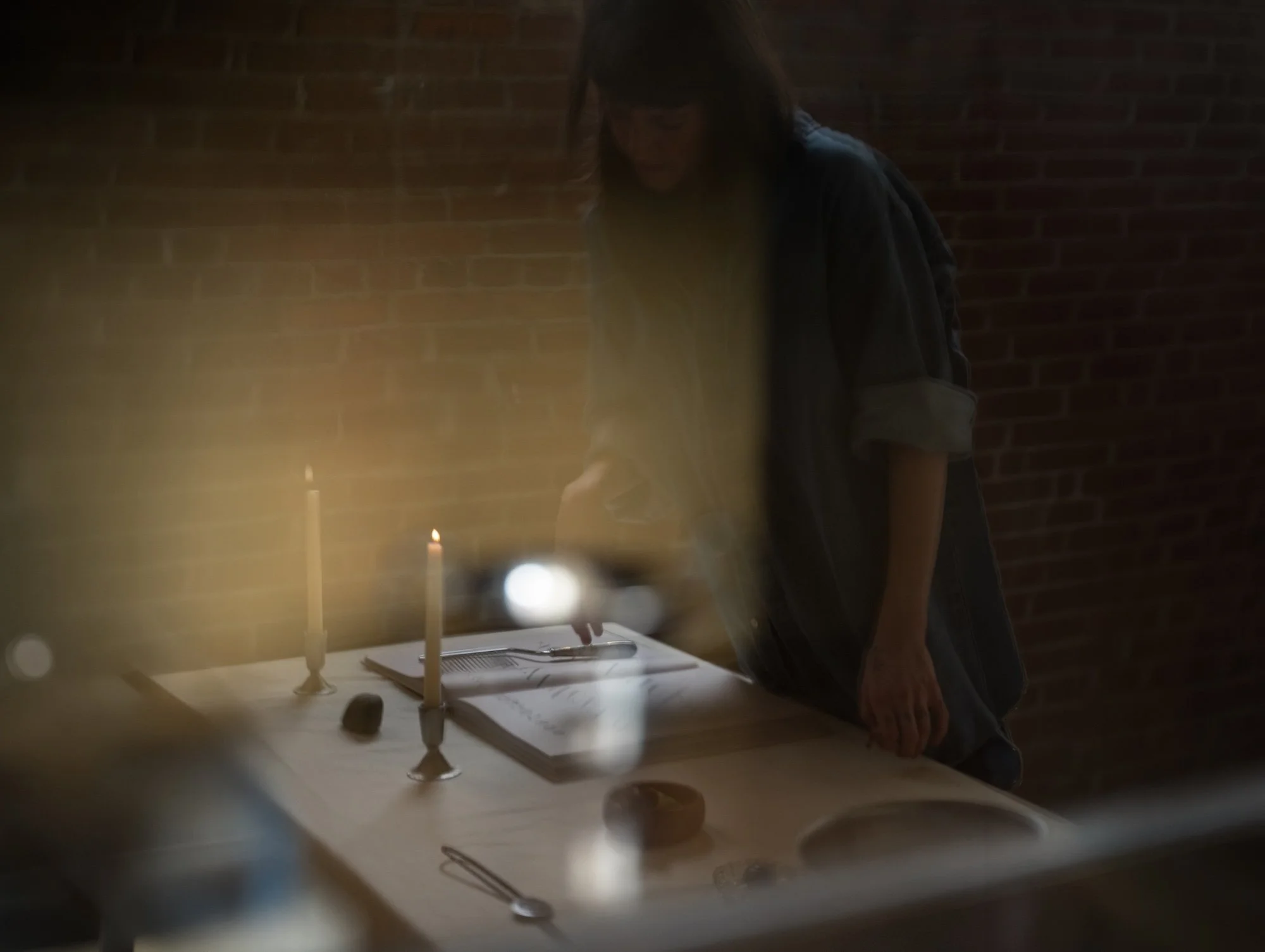 A person with dark hair leaning over a table illuminated by candlelight, with candles, a notebook, and utensils on the table, against a brick wall background.