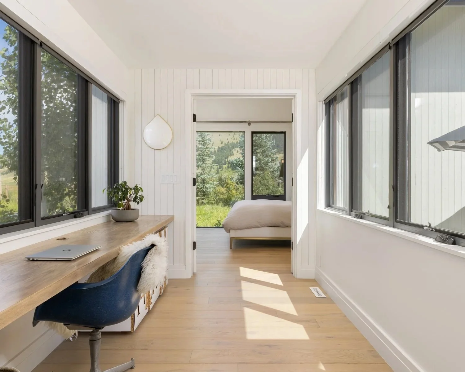 Room with large windows, wooden desk with laptop and plant, blue office chair, and door leading to bedroom with bed and scenic outdoor view
