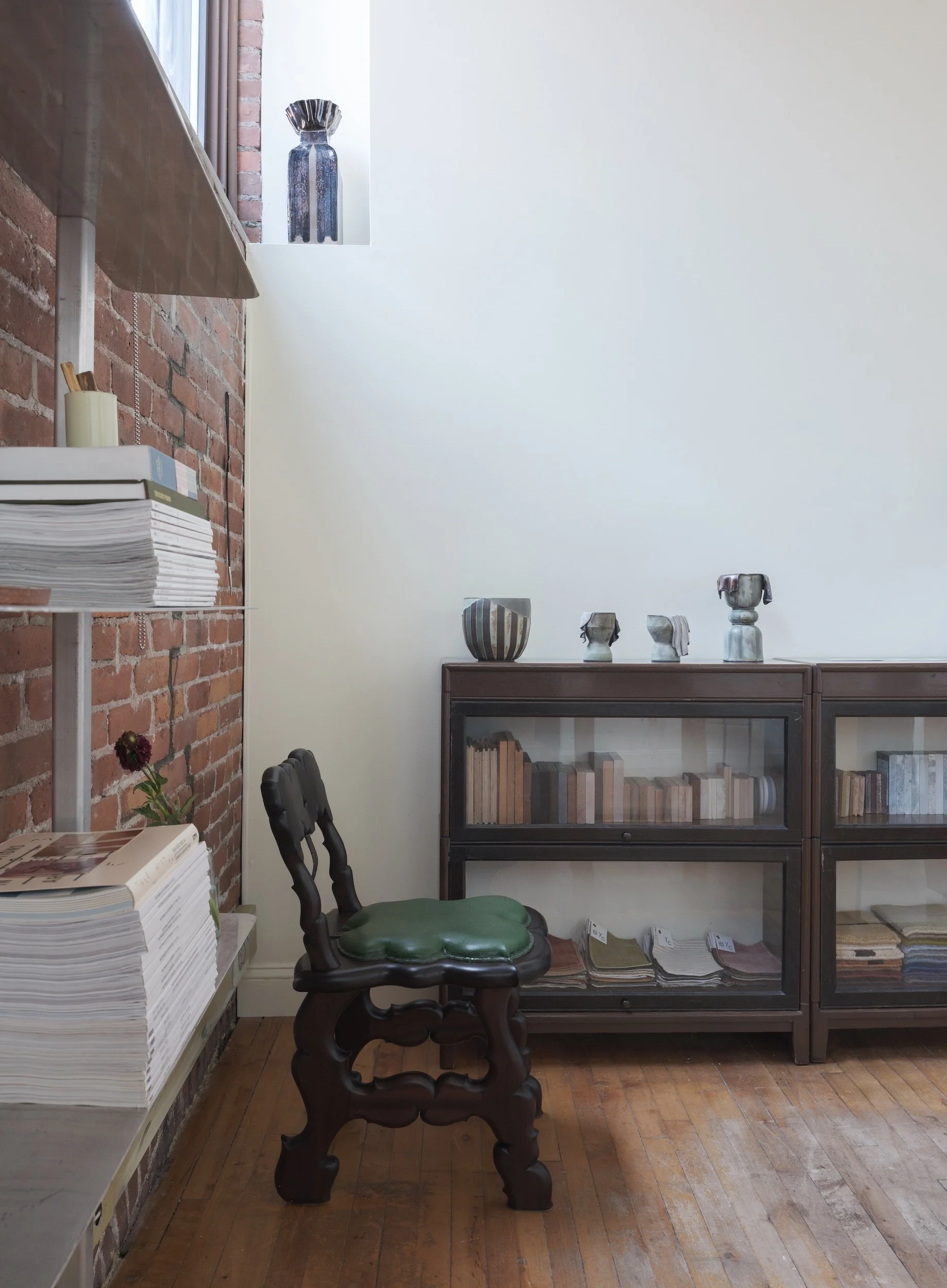 Interior of a room with wooden floors, an exposed brick wall, a metal shelving unit with stacks of magazines or papers, a flower in a pot, and a vintage wooden chair with a green cushioned seat next to two glass display cabinets with books and decorative pottery on top.