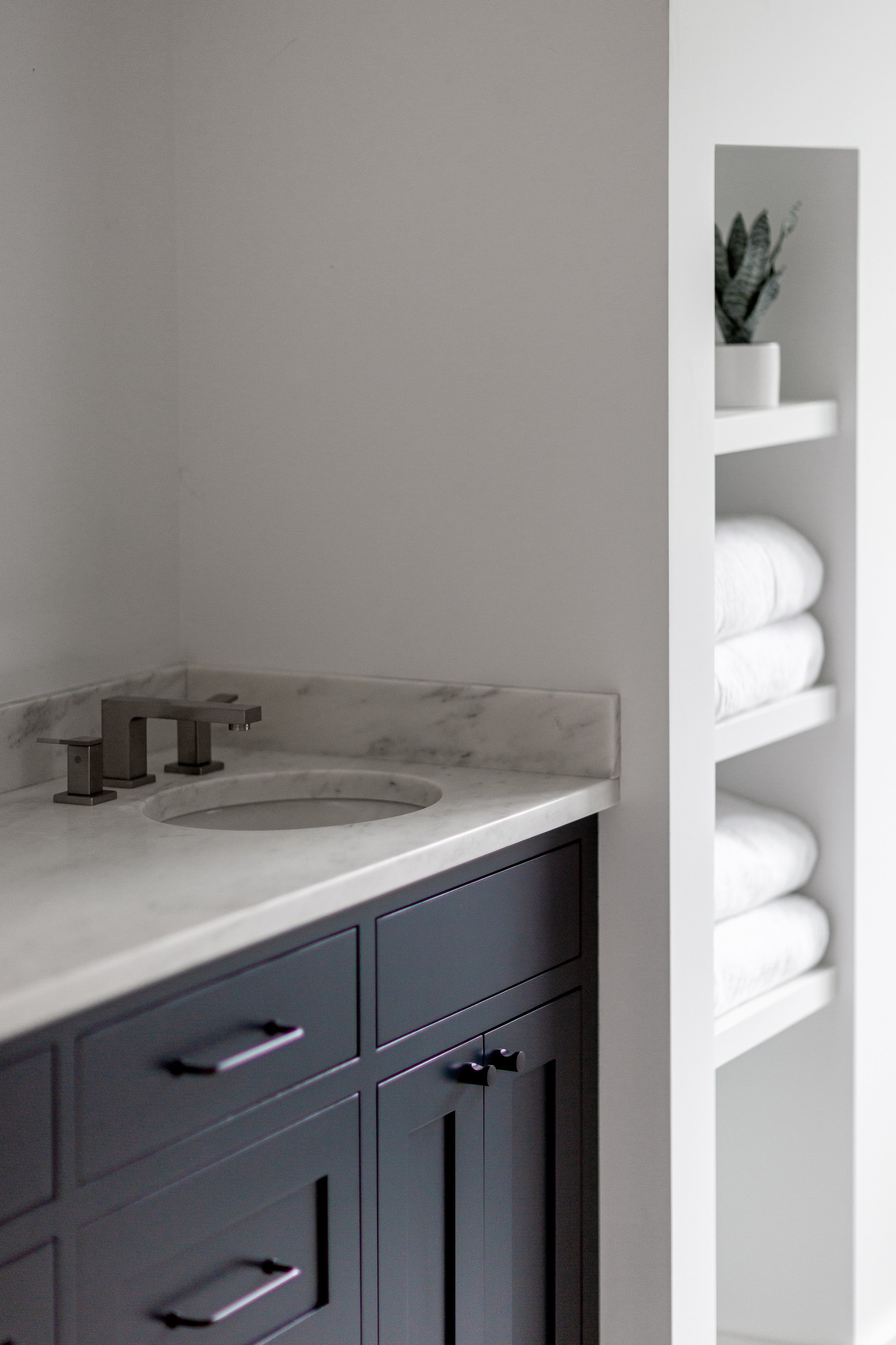 Modern bathroom with a white marble countertop, black cabinet with black handles, brushed metal faucet, and a built-in shelf with rolled white towels and a potted plant.