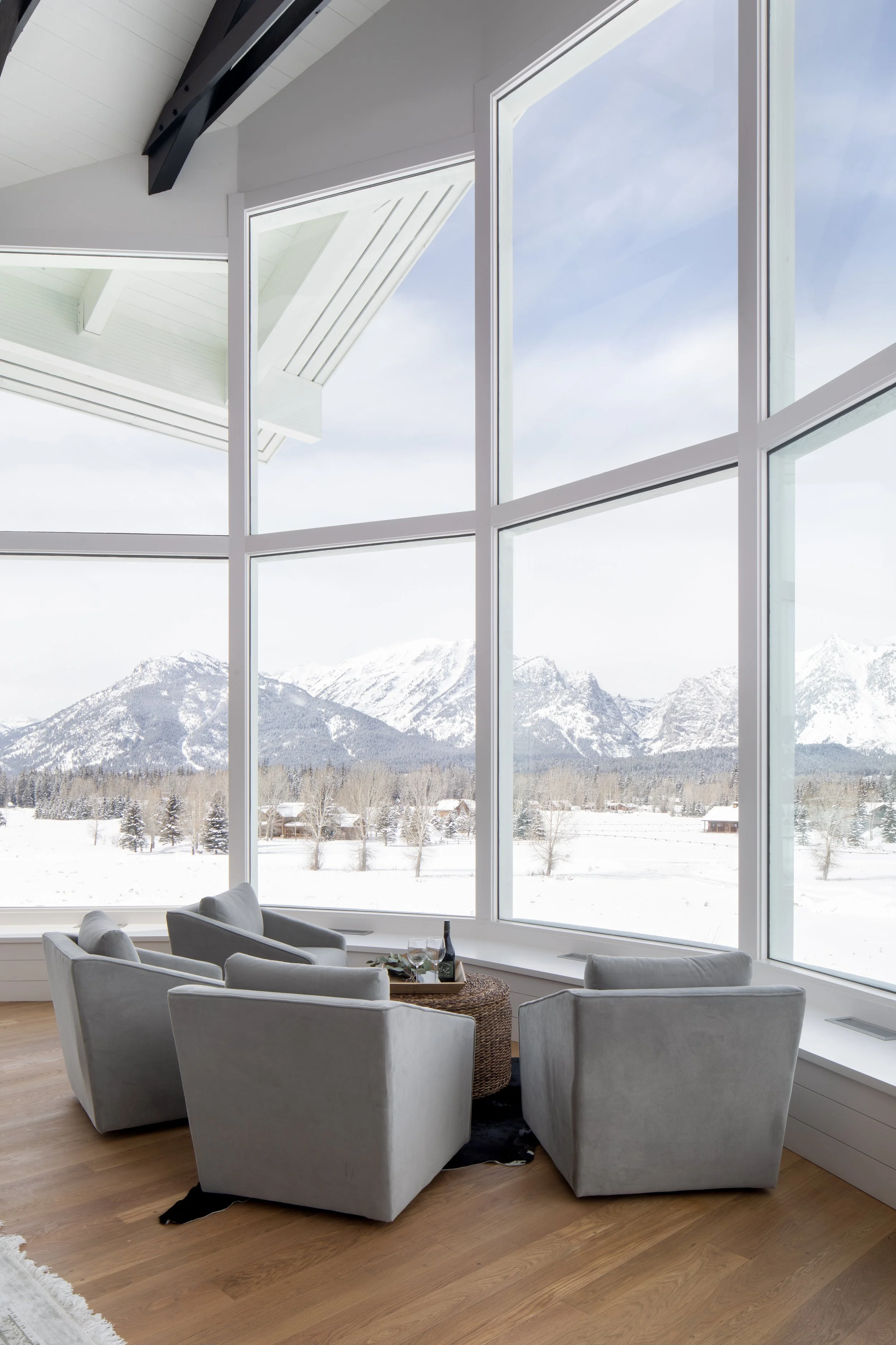 Interior of a modern living room with large floor-to-ceiling windows showing a snowy mountain landscape outside. There are four white armchairs arranged around a coffee table, with a bottle of wine and glasses on the table.