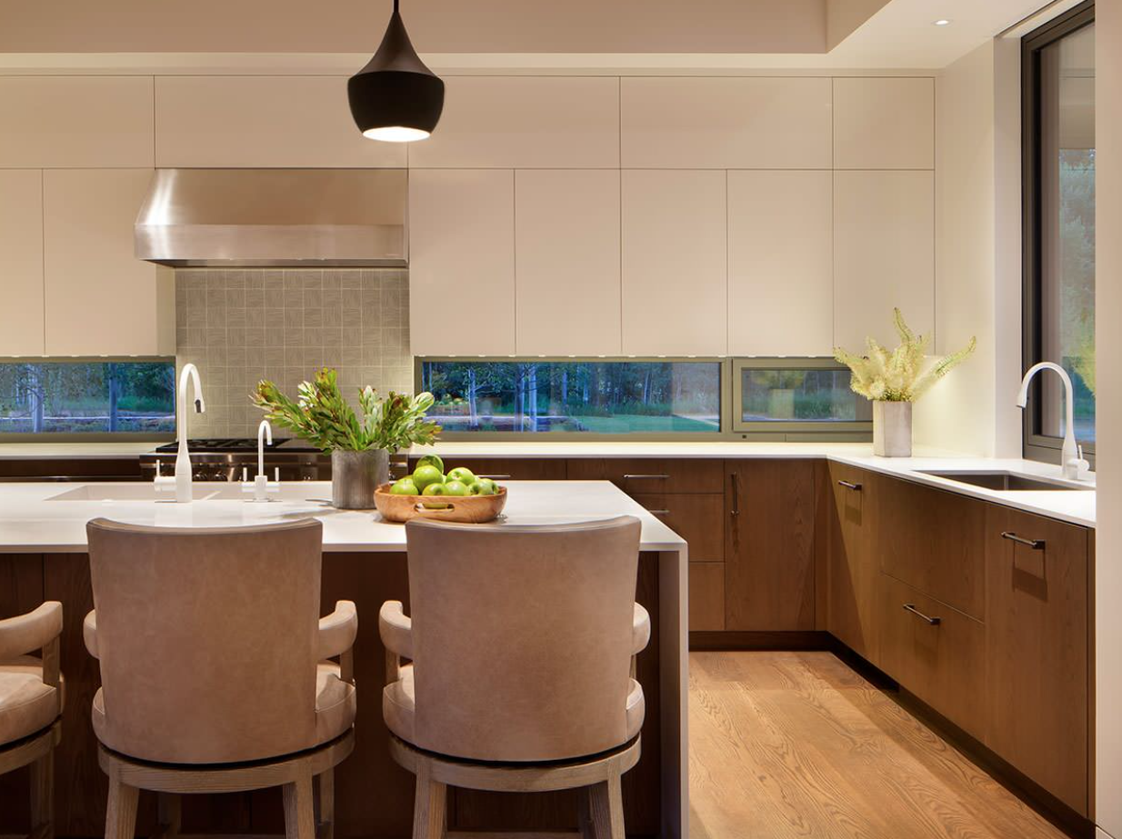 Modern kitchen with white and wood cabinets, a white island with a bowl of green apples and a plant, two chairs, stainless steel oven hood, and large windows showing outdoor trees.