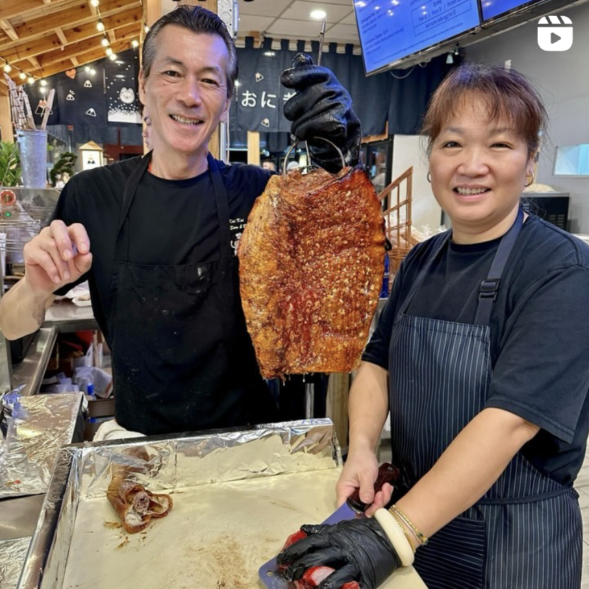 Two people in black shirts and aprons are smiling and holding a large, crispy roasted fish with a hook, in a kitchen or food stall. One person is holding the fish with a gloved hand, and the other is preparing to cut it. There are kitchen utensils, a cutting board, and a digital screen in the background.