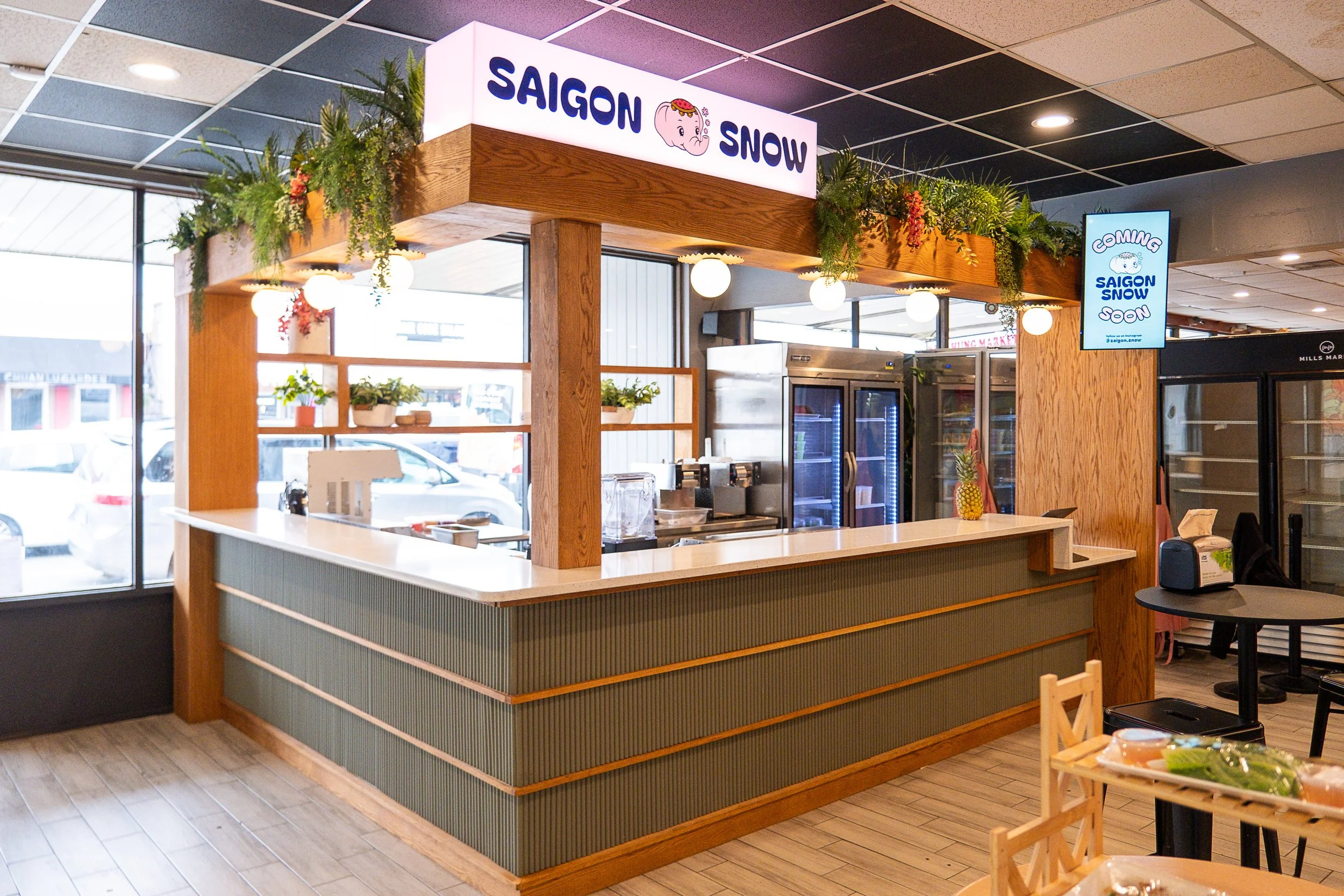A food service counter with a wooden frame, plants on top, and a sign that reads "Saigon Snow" inside a restaurant or cafe.