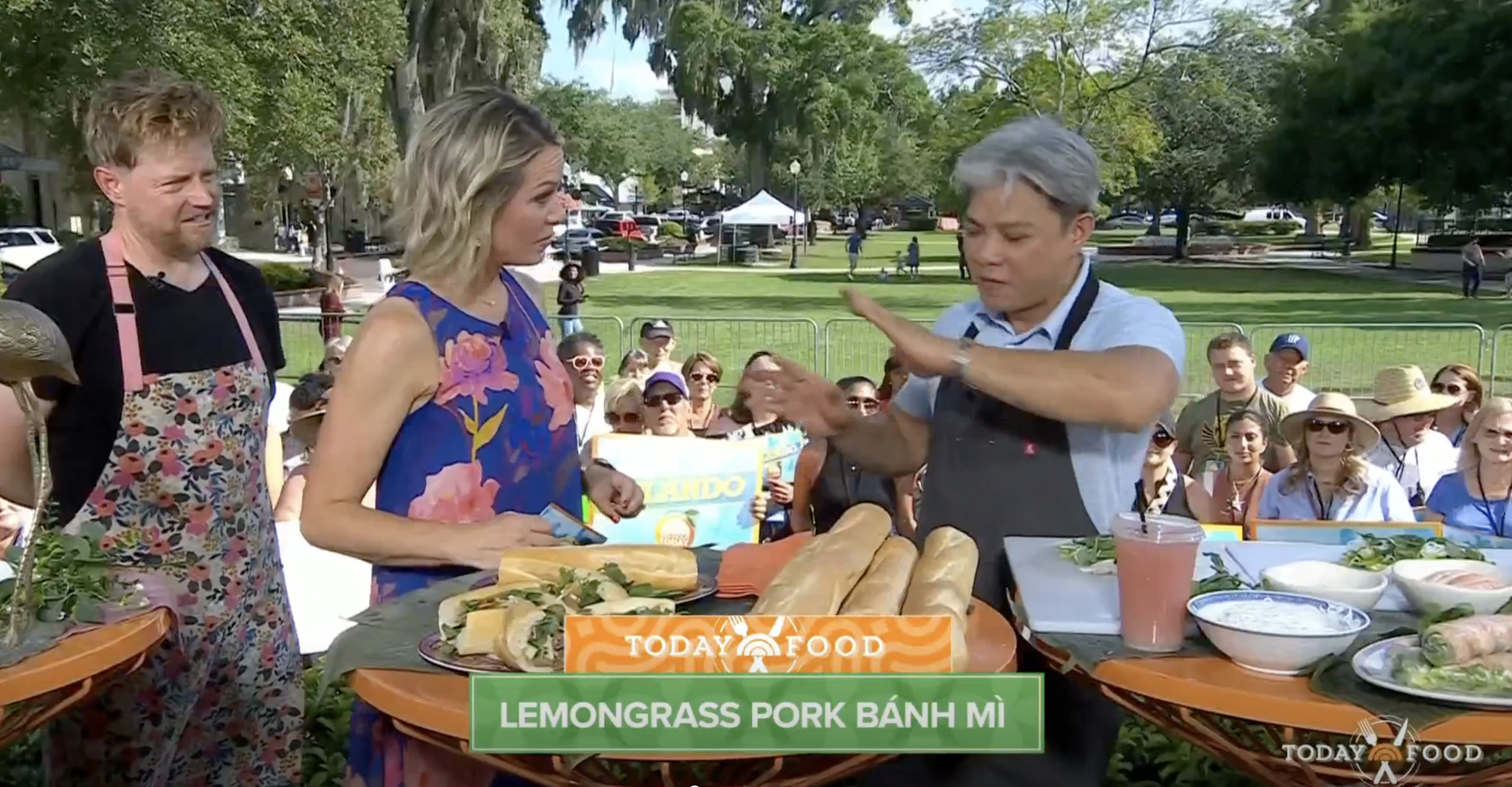 A chef demonstrating Thai-style pork banh mi sandwiches during a cooking segment at an outdoor event, with a woman host and a crowded park background.