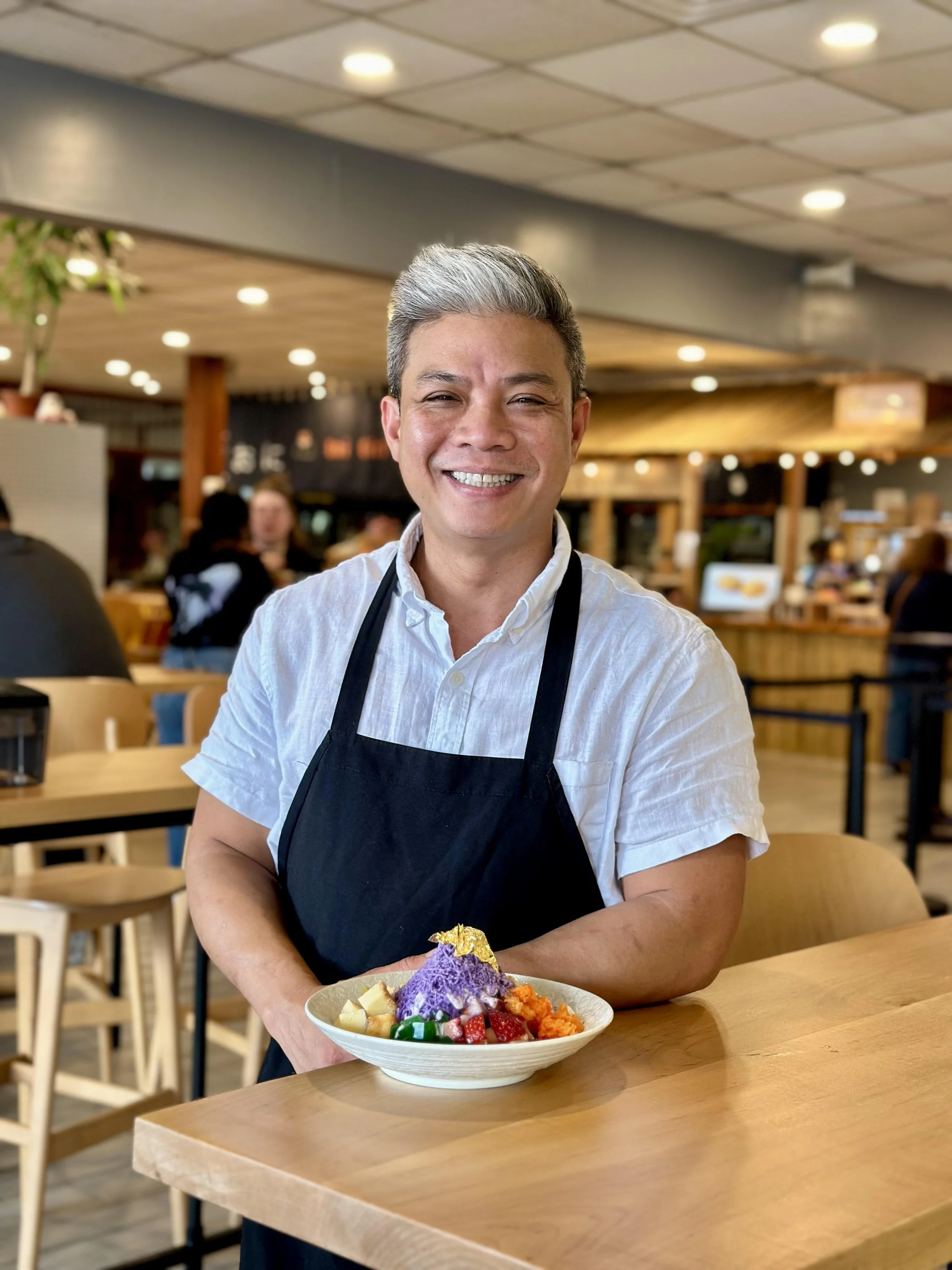 Smiling man in a white shirt and black apron holding a bowl of colorful shaved ice dessert inside a busy restaurant